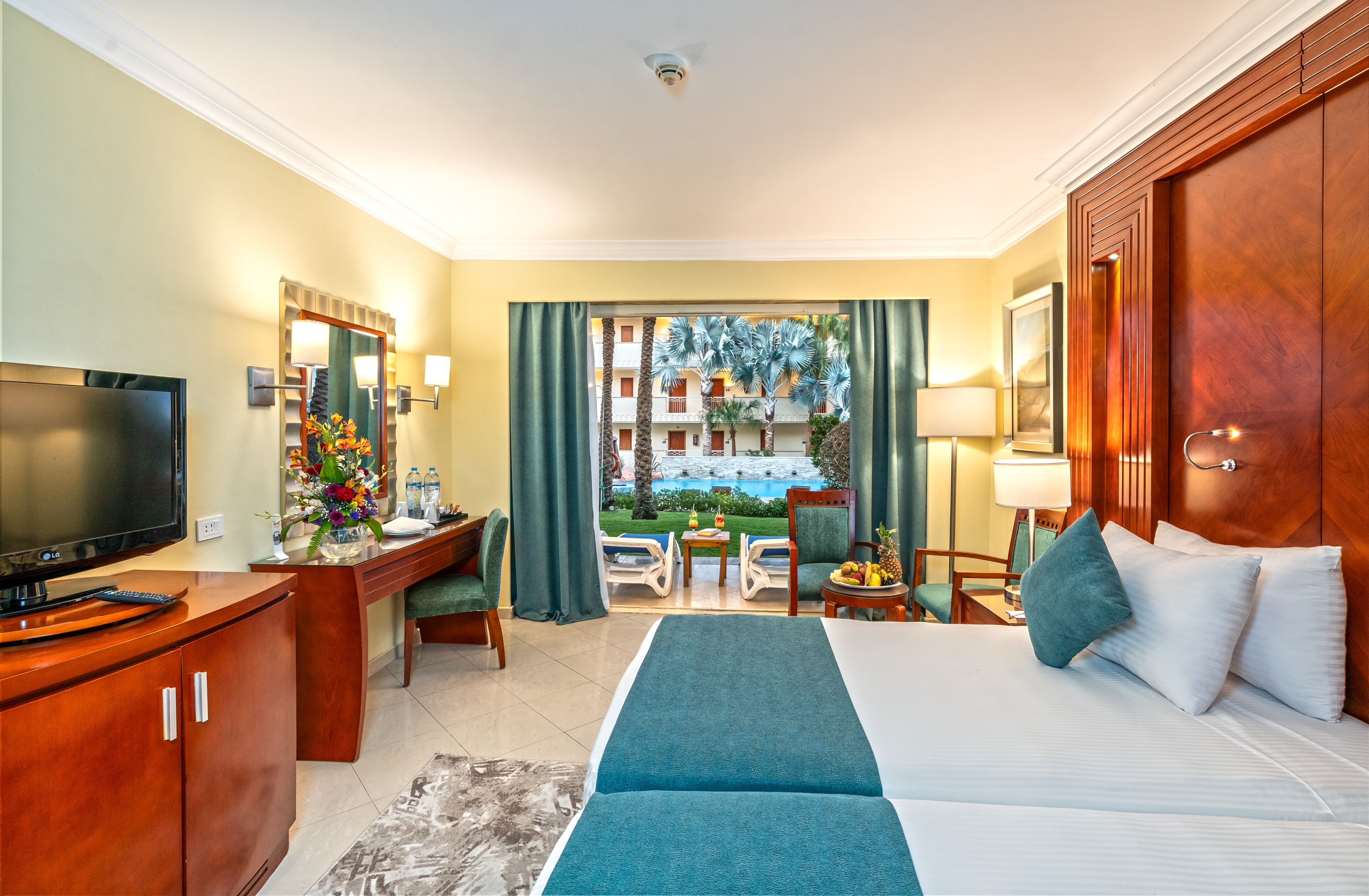 A hotel room with a bed, desk, TV, and a view of a pool and palm trees.