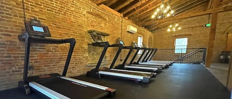 A gym with treadmills lined up against a brick wall, wooden beams on the ceiling, and a staircase leading to an upper level.