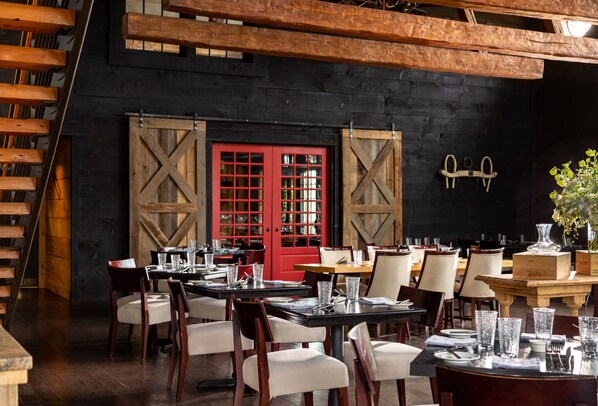 Un interior de restaurante con mesas y sillas de madera, puertas rojas y una pared negra con vigas de madera.