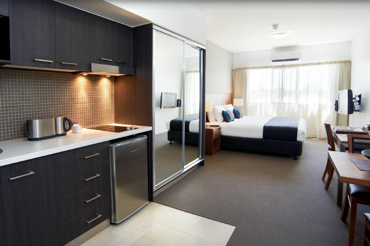 A modern hotel room with a kitchenette, a bed, a desk, and a TV.