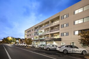 Hotel a più piani dal design moderno, con balconi su ogni piano, un parcheggio con diverse auto e un cielo sereno.