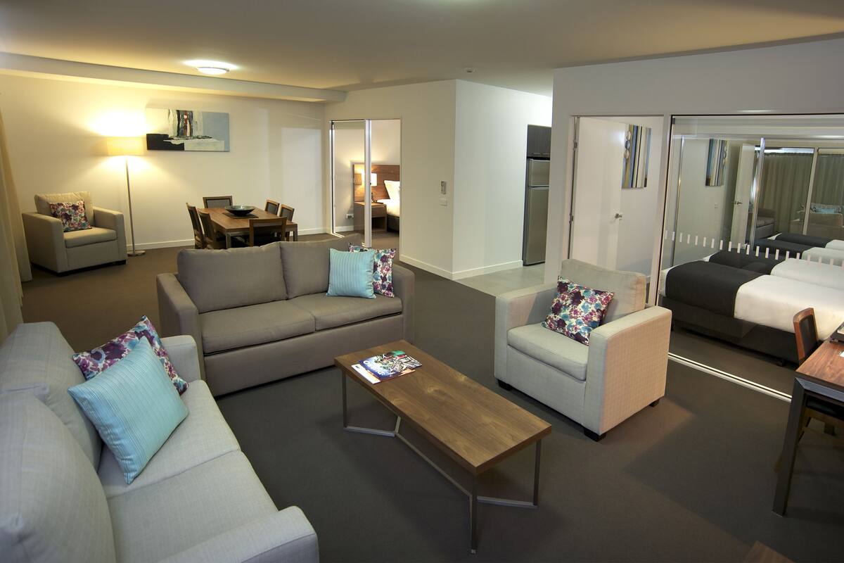 A modern living room with a sofa, armchair, and a coffee table. There is a dining table with chairs and a refrigerator in the background.