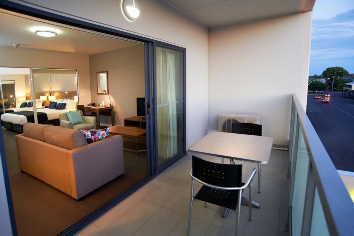 A modern hotel room with a balcony, a sofa, a dining table, and a small kitchen area.