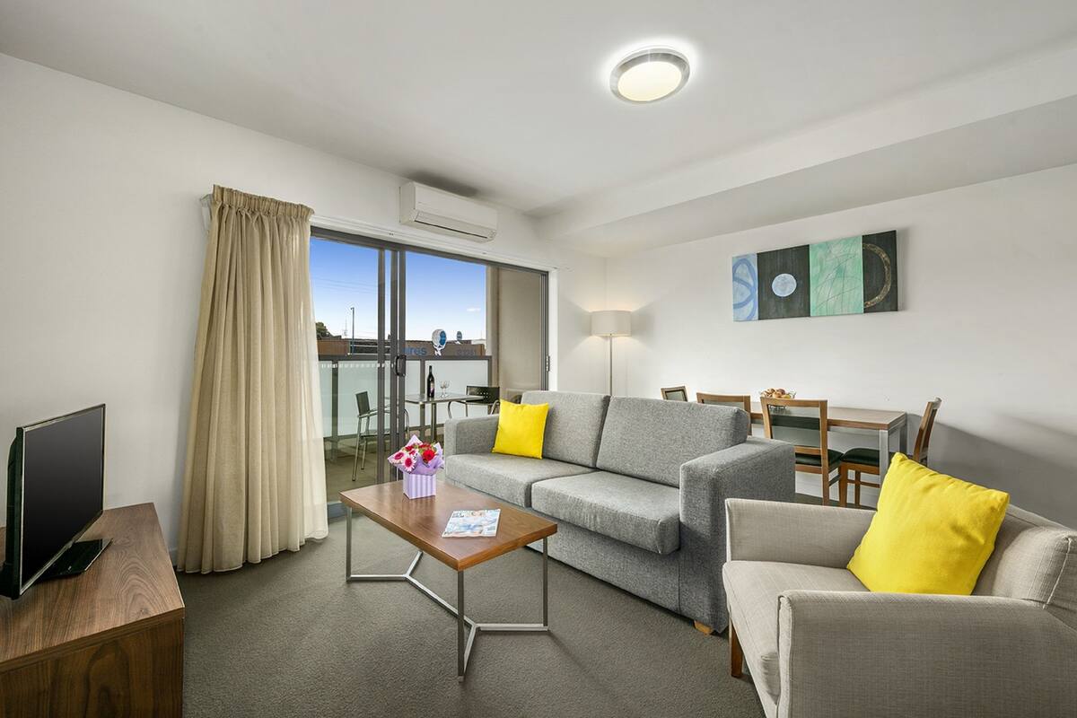A modern hotel room with a grey sofa, yellow cushions, a wooden coffee table, a flat-screen TV, and a dining area with a table and chairs.
