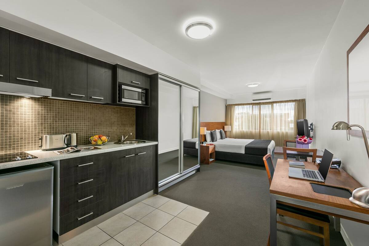 A modern hotel room with a kitchenette, a bed, a desk with a laptop, and a bedside table with a lamp.