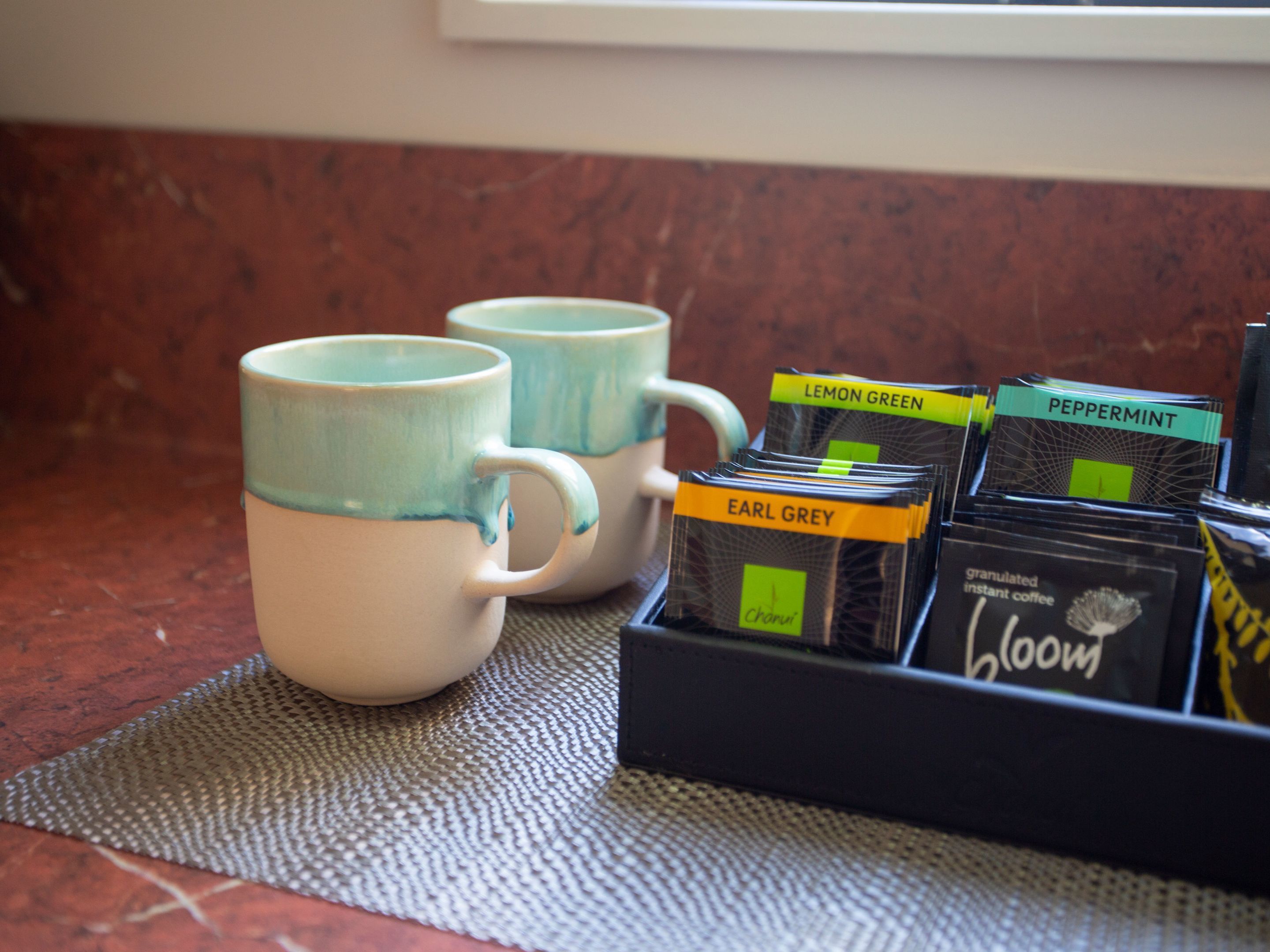 Zwei Keramiktassen mit blauer Glasur und eine Schale mit Instantkaffeepäckchen, beschriftet mit Sorten wie Zitrone-Grün, Pfefferminze und Earl Grey.