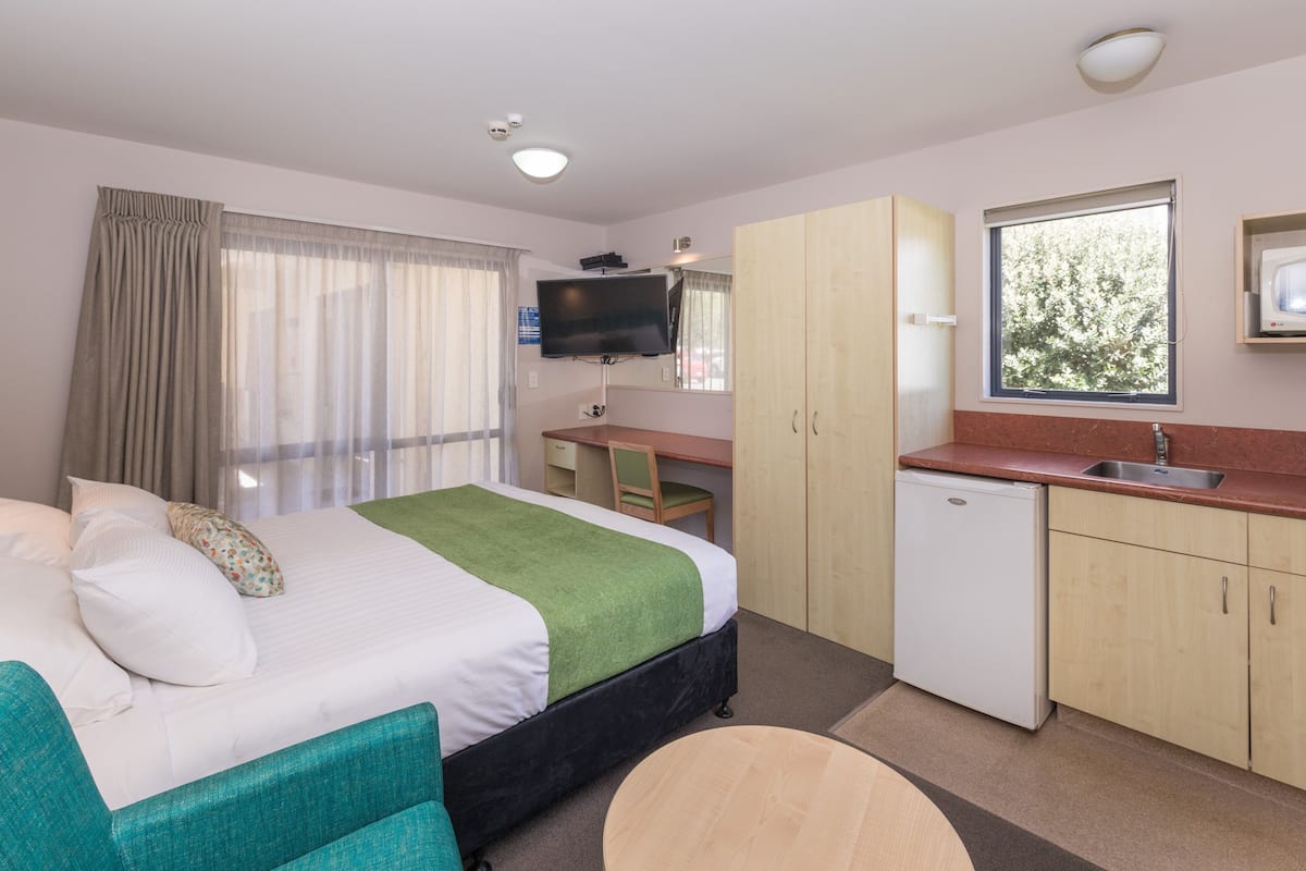A hotel room with a bed, a desk, a television, a small refrigerator, and a window with a view of trees.