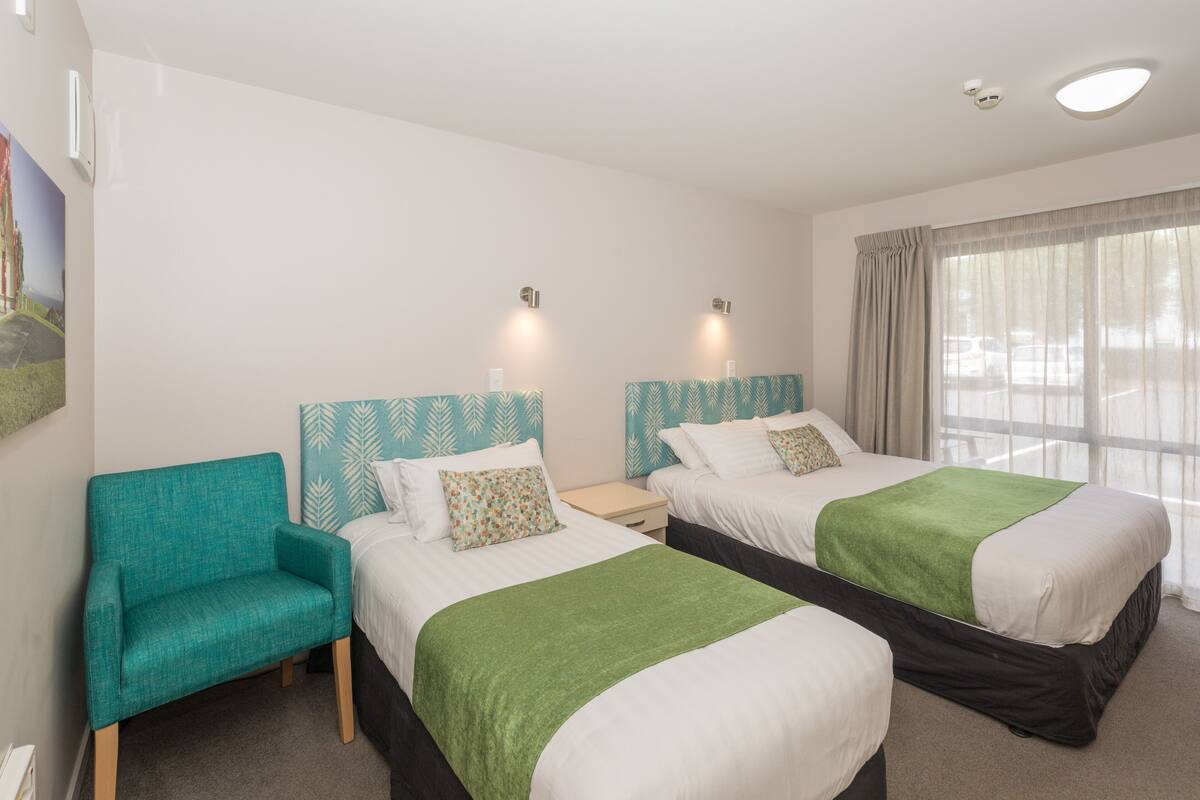 A hotel room with two beds, a teal armchair, and a window with curtains.