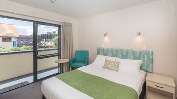 A hotel room with a large bed, a green throw, a teal armchair, a small table, and a view of the outside.