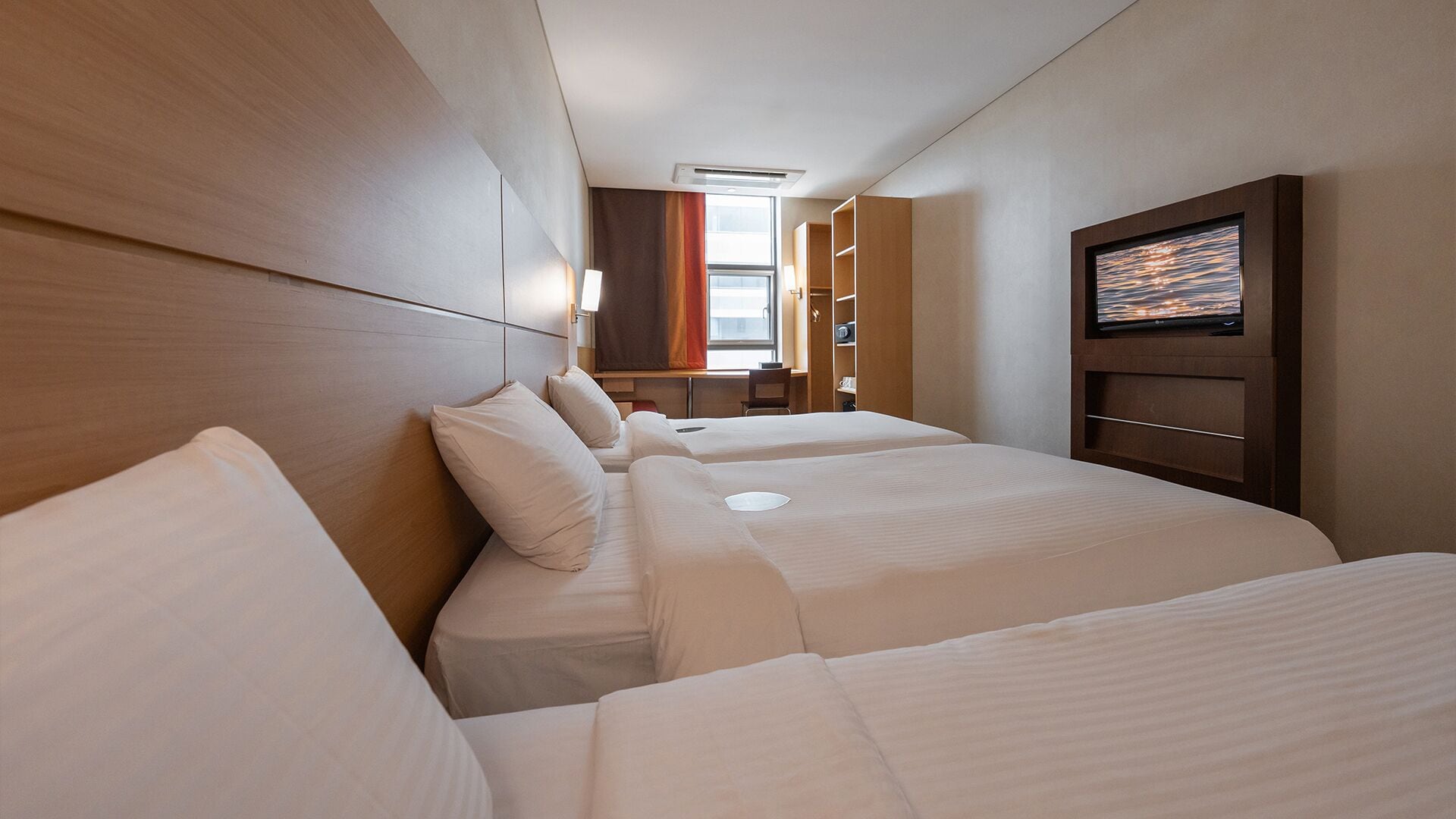 A hotel room with two beds, a desk, a chair, a TV, and a window.