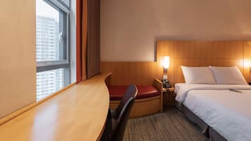 A hotel room with a desk, a chair, a bed, a nightstand, and a window with a view of buildings.