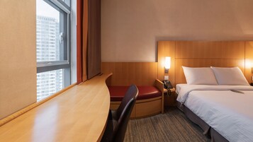 Habitación de hotel con escritorio, una silla, cama, mesita de noche y ventana con vista a edificios.