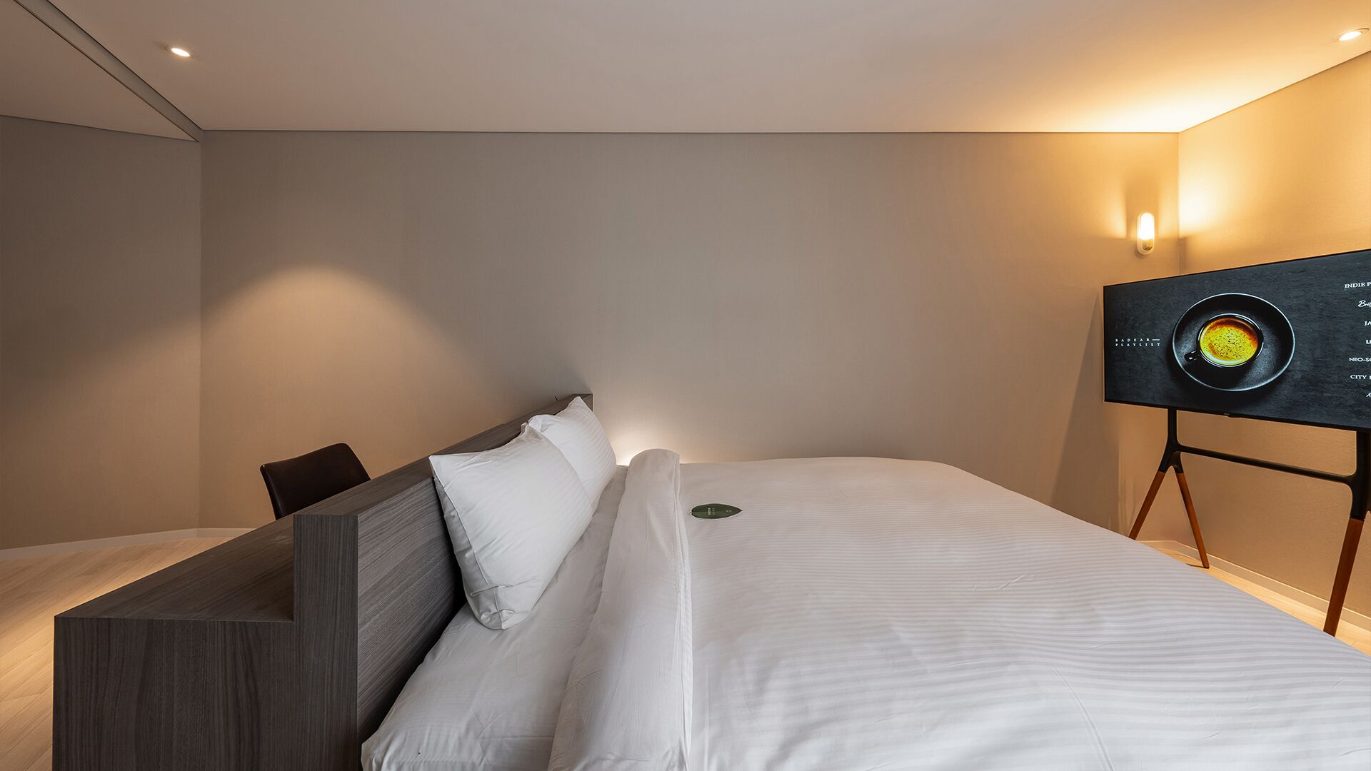 A modern hotel room with a bed, a seating area, and a wall-mounted screen displaying a menu.