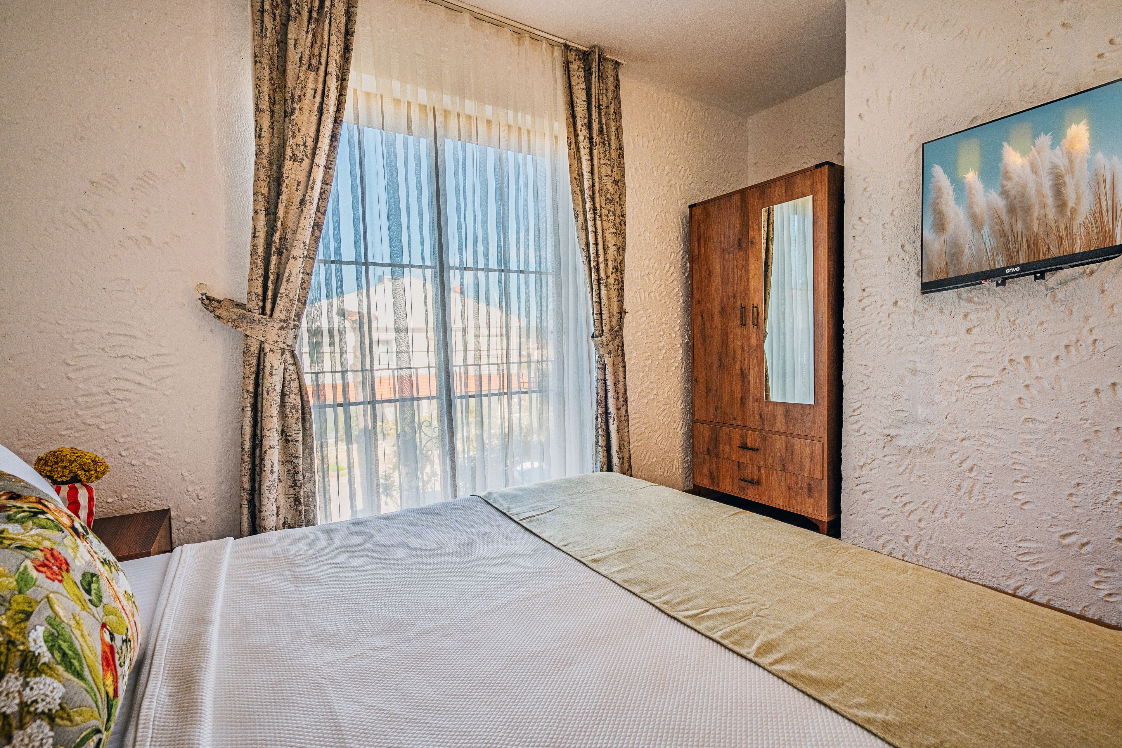 A bedroom with a bed, curtains, a wooden wardrobe, and a television on the wall.
