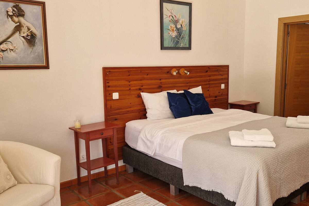 Ein Schlafzimmer mit Holz-Kopfteil, einem Bett mit weißer und blauer Bettwäsche, einem kleinen Nachttisch und einem Sessel.