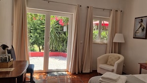 Une chambre d’hôtel avec un lit, une chaise, un bureau avec une lampe, une petite table et une fenêtre avec des rideaux.
