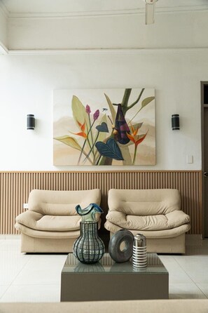 A modern living room with two beige sofas, a glass coffee table, and a wall-mounted painting of a plant.