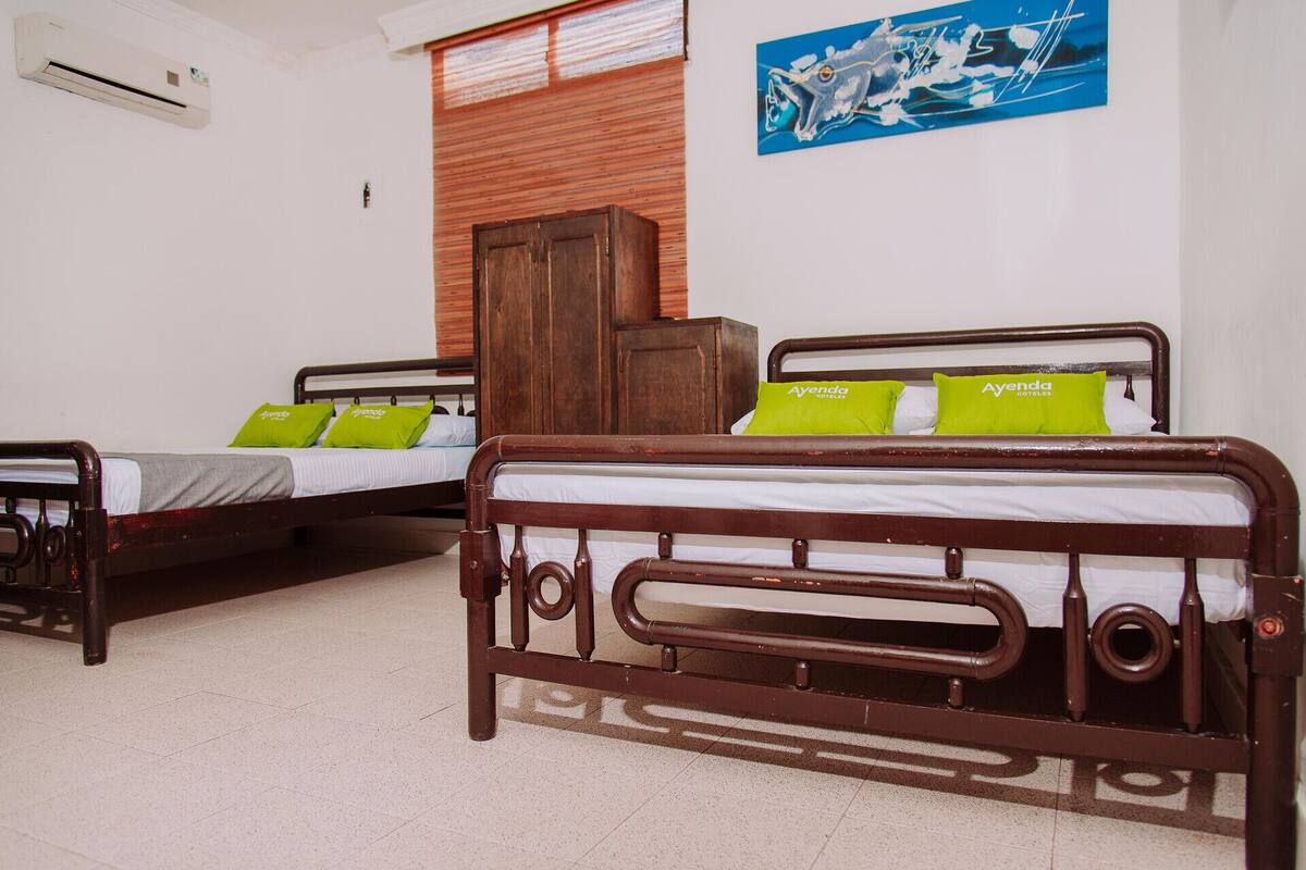 Une chambre d’hôtel avec deux lits en bois, chacun agrémenté d’oreillers verts et d’une tête de lit en bois. On y trouve également une armoire en bois et un tableau bleu accroché au mur.