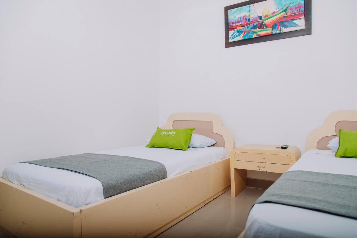 Une chambre double avec deux lits, chacun doté d’un cadre en bois et d’un oreiller vert, ainsi qu’une petite table de chevet et un tableau accroché au mur.
