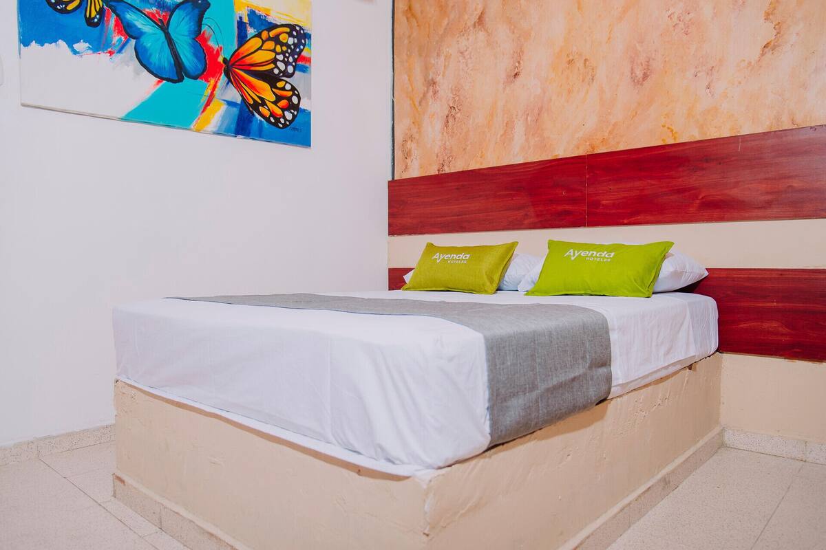Une chambre d’hôtel avec un lit, deux oreillers aux couleurs de la marque « Ayenda Hoteles », un tableau coloré accroché au mur et un mur texturé derrière le lit.