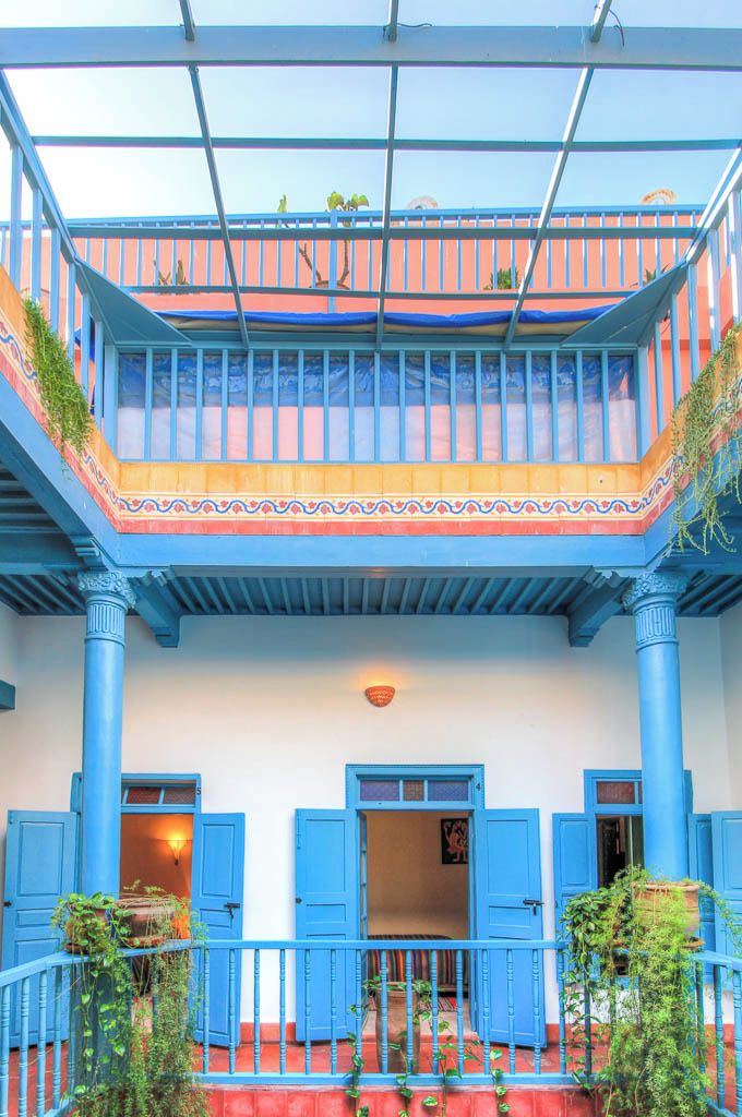 Un edificio de dos pisos con puertas azules y columnas, un balcón con plantas y un primer piso de color rojo.