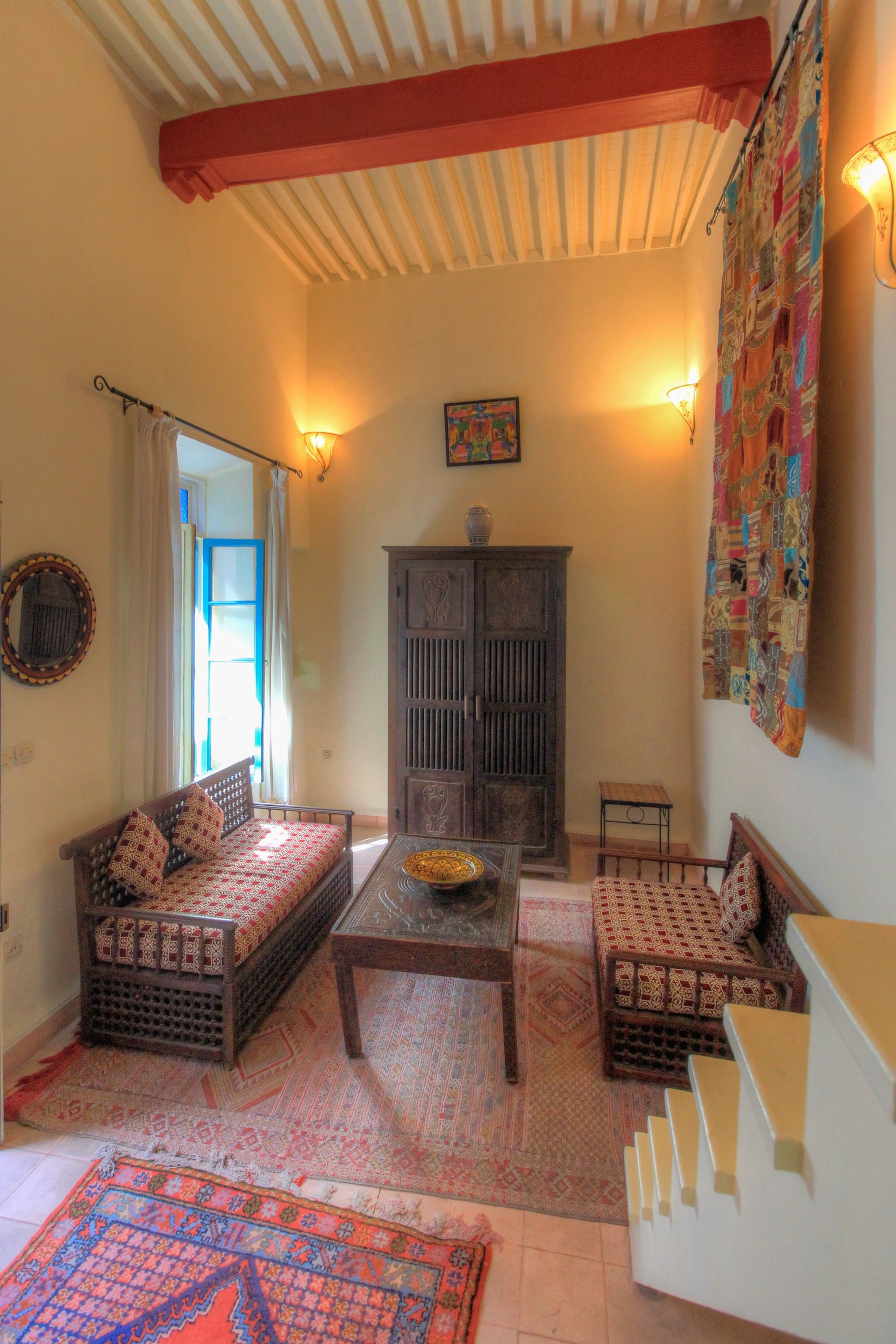 A cozy living room with a sofa, a coffee table, and a cabinet. There is a patterned rug on the floor, and a colorful curtain hangs on the wall.