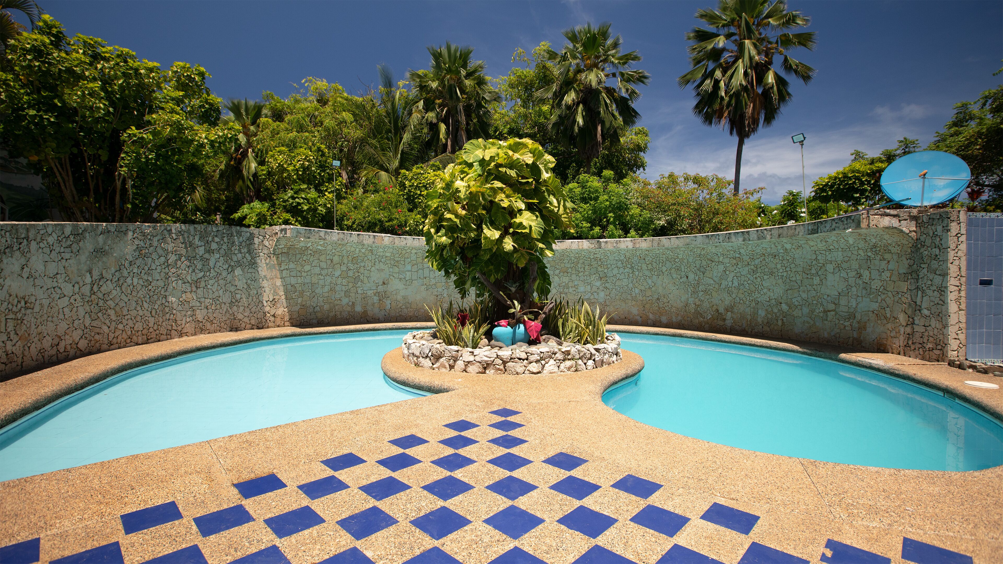 Una piscina con un diseño de ajedrez en el suelo, rodeada por un muro de piedra y palmeras.