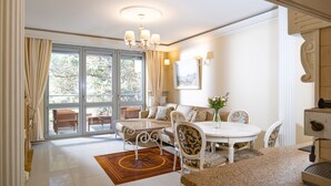 A well-lit room with a dining table, chairs, and a sofa. There is a large window with curtains, a chandelier, and a painting on the wall. - Dariusz Apartament PREMIUM Jurata z garażem (Jurata)