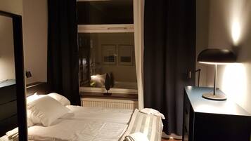 A neatly made bed with white linens, a bedside table with a lamp, and a view of a room with a window and curtains.