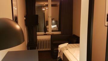 Habitación de hotel pequeña con cama, escritorio, una silla y una mesita. Cuenta con ventana con cortinas, radiador y lámpara colgante.