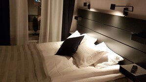 A neatly made bed with white linens and a black pillow, a bedside table with a remote control, and a window with curtains partially drawn. - Hotell Kong Christian (Kristianstad)