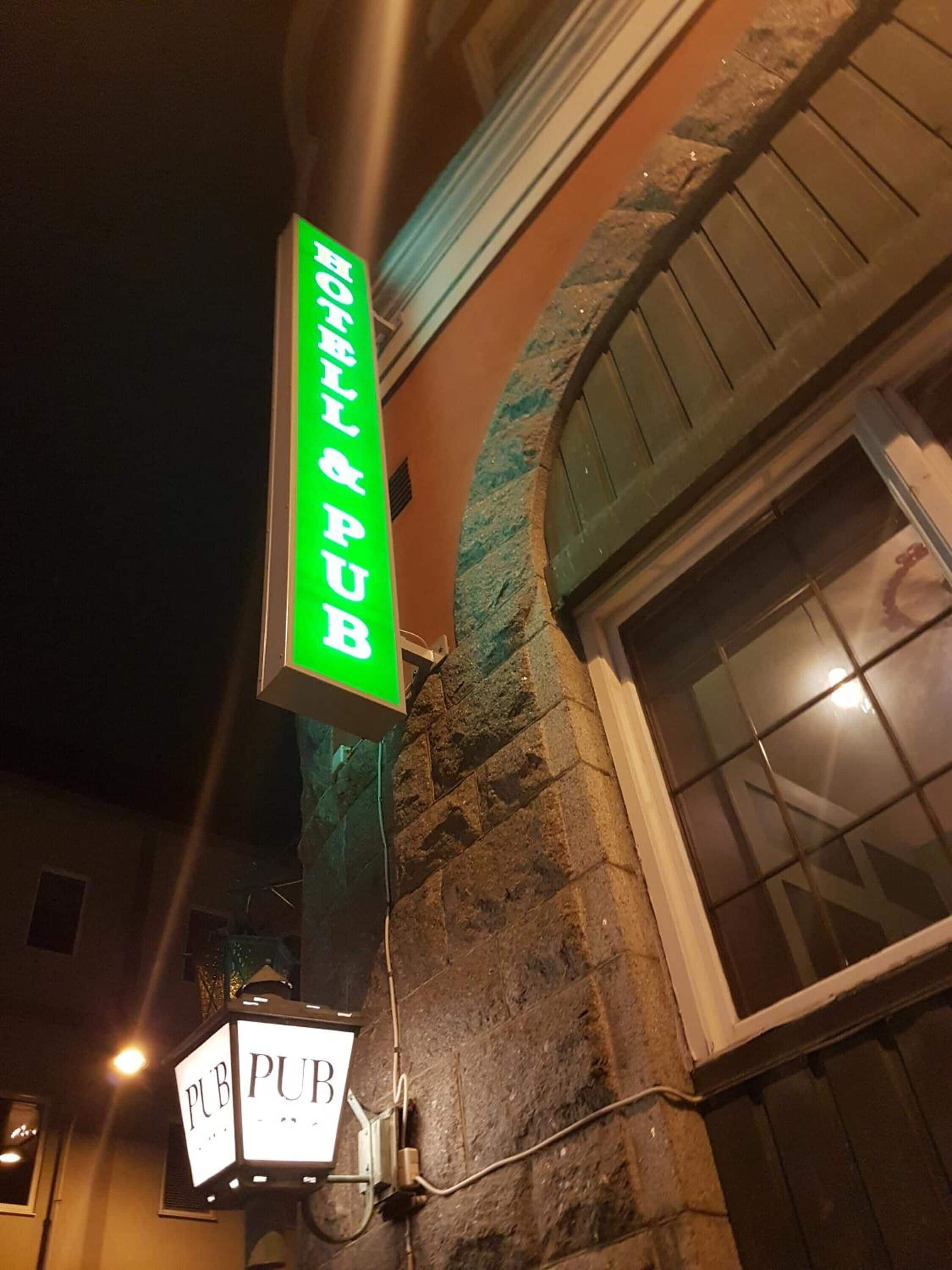Un letrero de neón para un pub con el nombre 'ROBB'S PUB' en letras blancas sobre un fondo verde, sujeto a una pared de piedra.