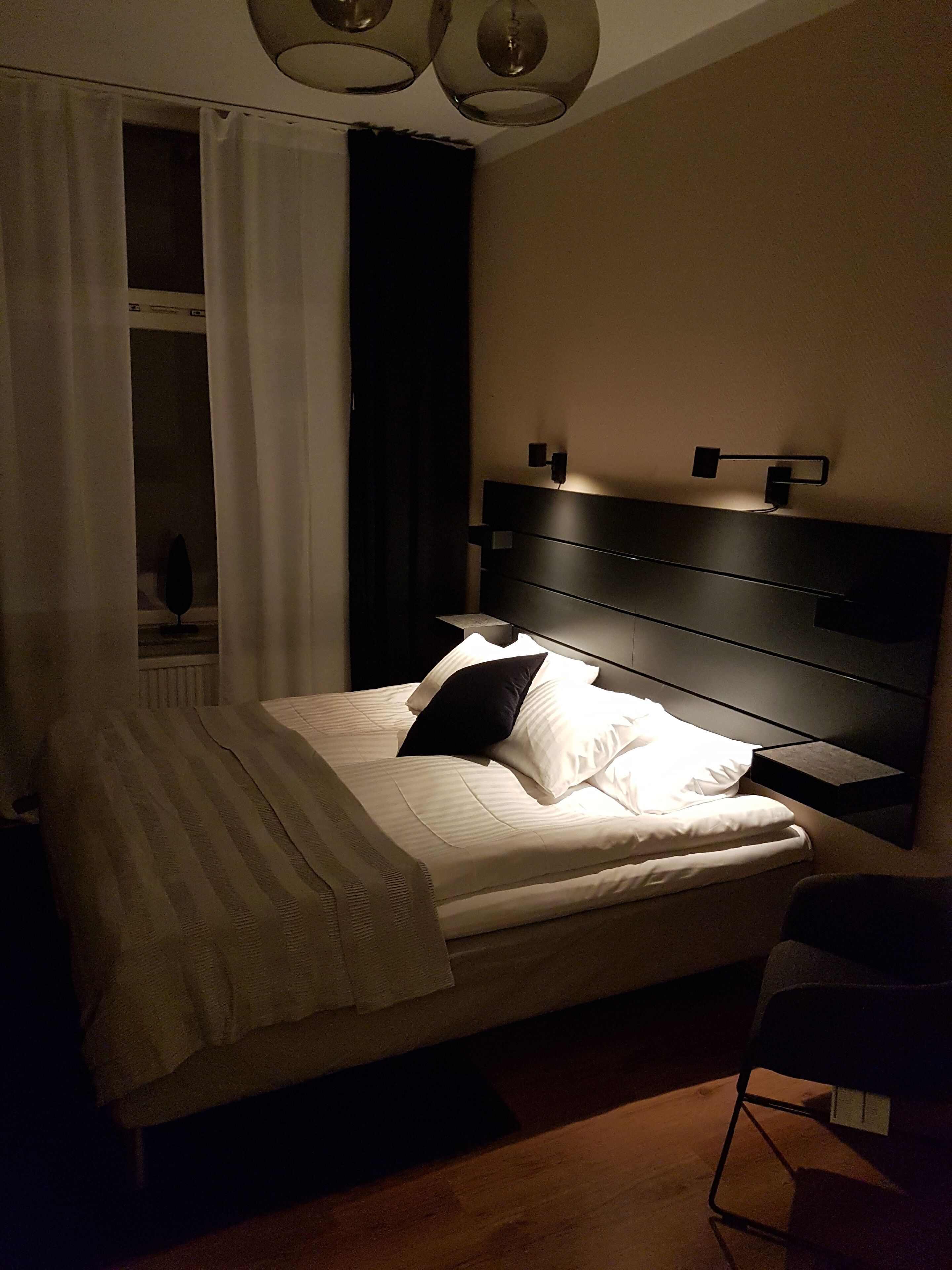 Una cama bien hecha con ropa blanca y una almohada negra, una mesita de noche y una lámpara de pared.