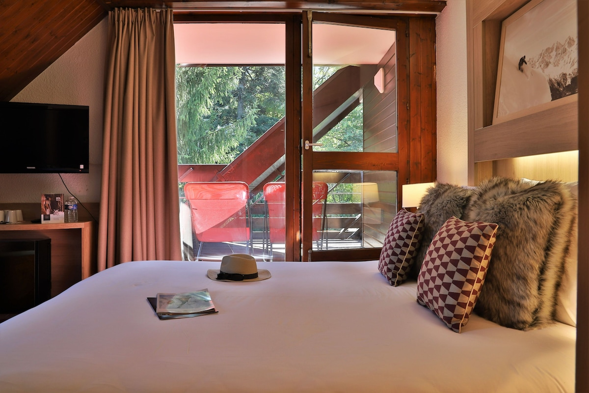A hotel room with a bed, a television, a window with curtains, and a view of an outdoor area with a chair and a staircase.