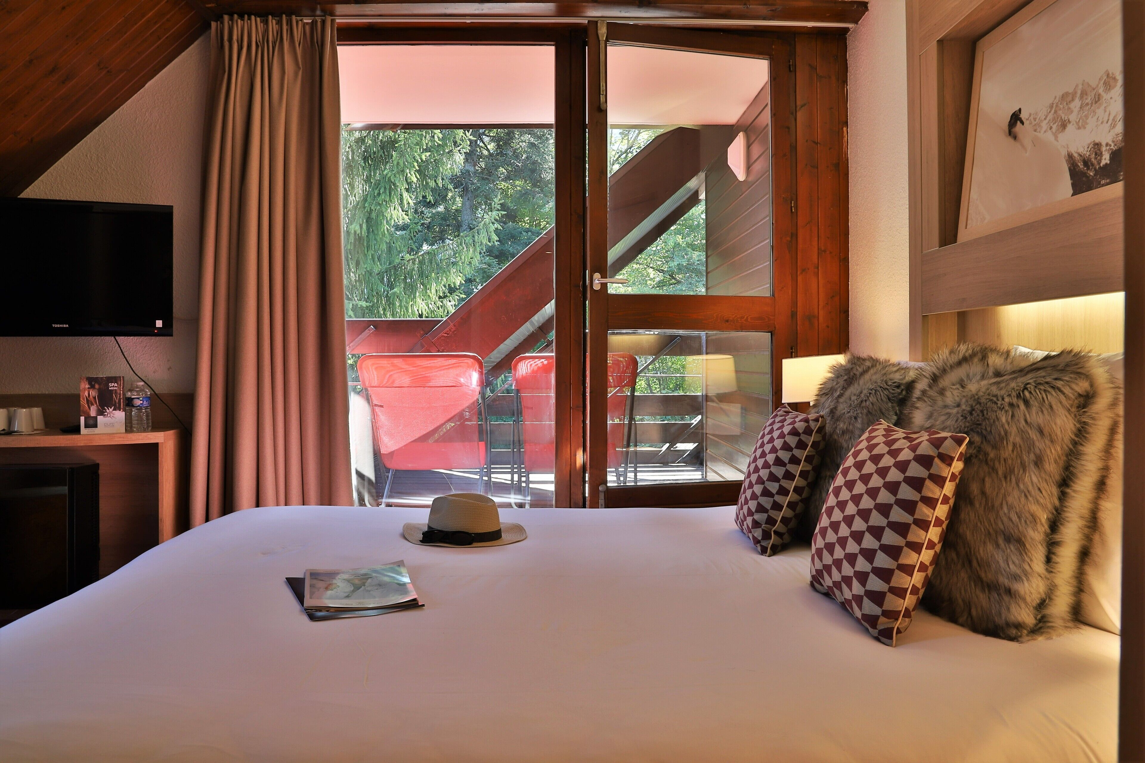 A hotel room with a bed, a television, a window with curtains, and a view of an outdoor area with a chair and a staircase.