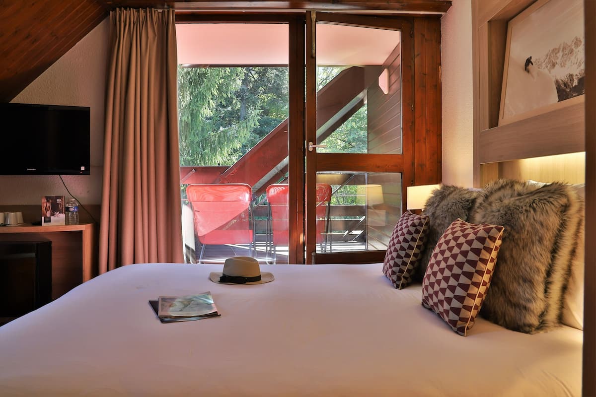 A hotel room with a bed, a television, a window with curtains, and a view of an outdoor area with a chair and a staircase.