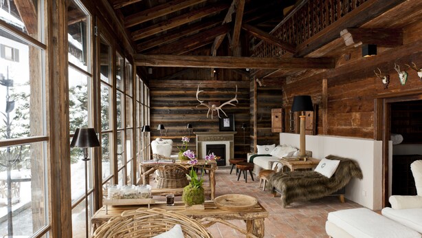 A rustic log cabin interior with large windows, a fireplace, wicker furniture, and a fur-covered chair. - House of Hütter - Wachtelhof Hotel & Spa (Maria Alm am Steinernen Meer)