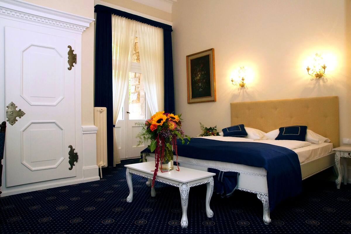 Een hotelkamer met een bed, nachtkastje, een tafel met bloemenvaas, een deur en een raam met gordijnen.