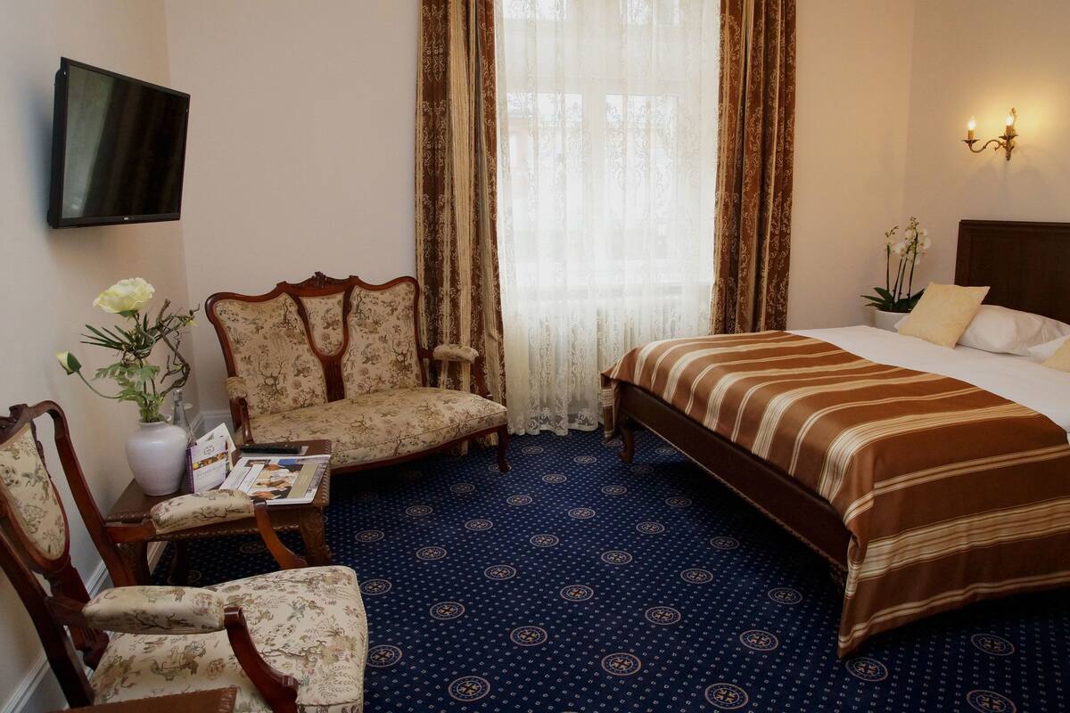 Hotelkamer met een bed, twee stoelen, een kleine tafel met een vaas en boeken, een aan de muur bevestigde televisie en een raam met gordijnen.