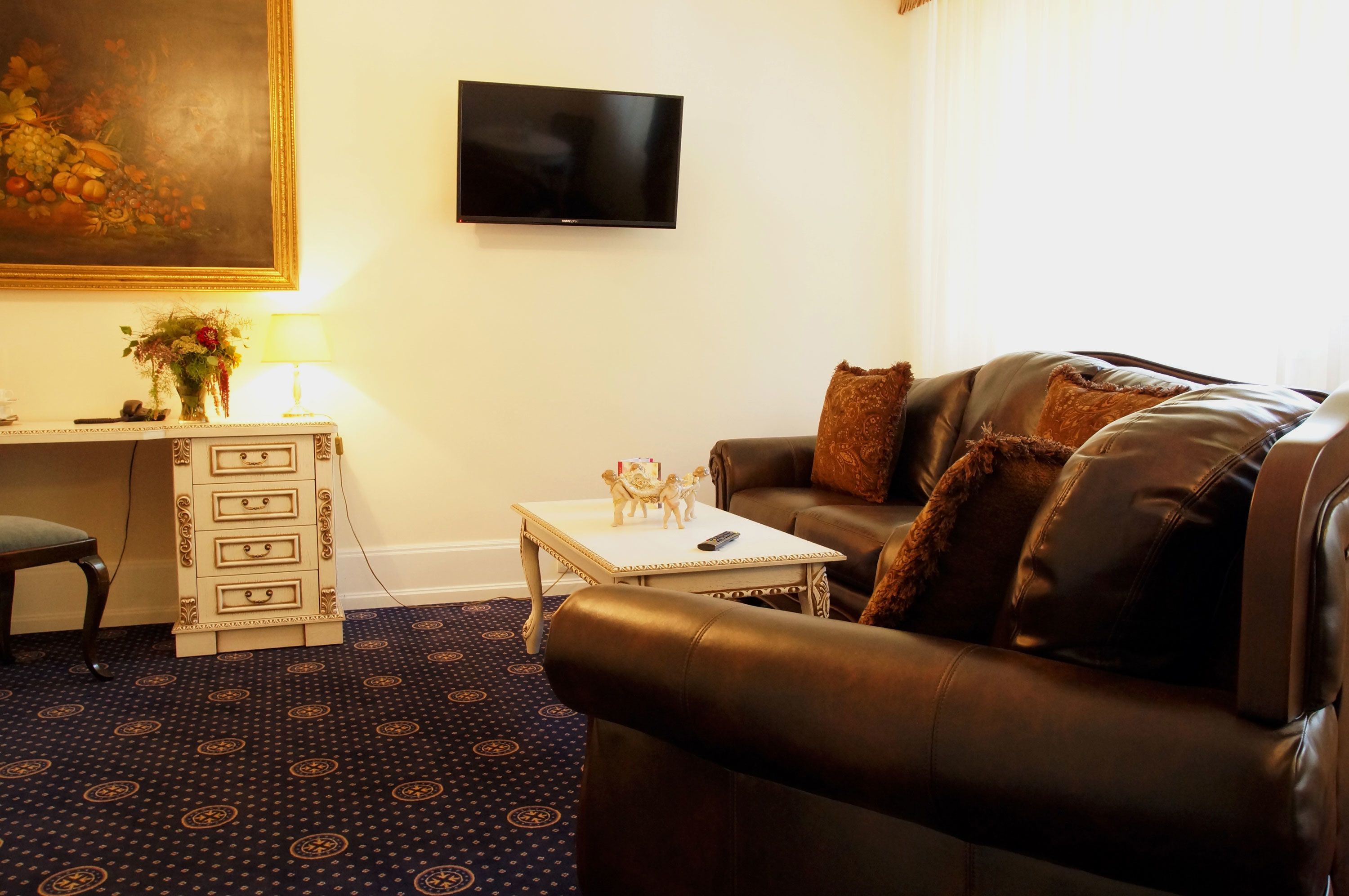 A living room with a brown leather sofa, a small white table with a lamp, a TV mounted on the wall, and a patterned carpet.