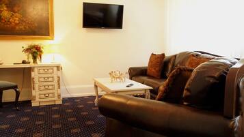 A living room with a brown leather sofa, a small white table with a lamp, a TV mounted on the wall, and a patterned carpet.
