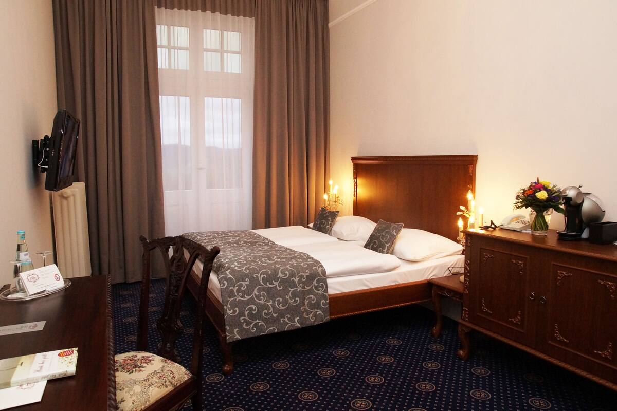 Een hotelkamer met een opgemaakt bed, een houten dressoir, een bureau met stoel, en een raam met gordijnen.