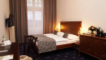 A hotel room with a neatly made bed, a wooden dresser, a desk with a chair, and a window with curtains.