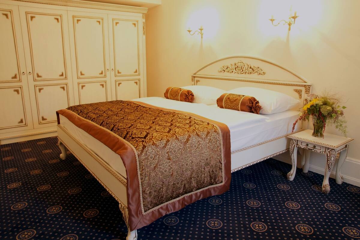 Een net opgemaakt bed met een beddekleed met een patroon en kussens, een klein nachtkastje met een bloemenvaas, en een grote garderobe met meerdere deuren.