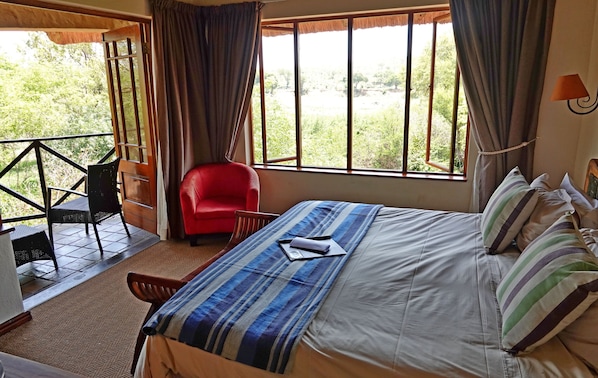 Habitación de hotel con cama, una silla roja, balcón con barandilla y vista a zonas verdes.