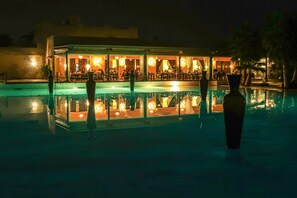 Vista nocturna de un hotel con piscina, mostrando un interior bien iluminado y un reflejo en el agua.