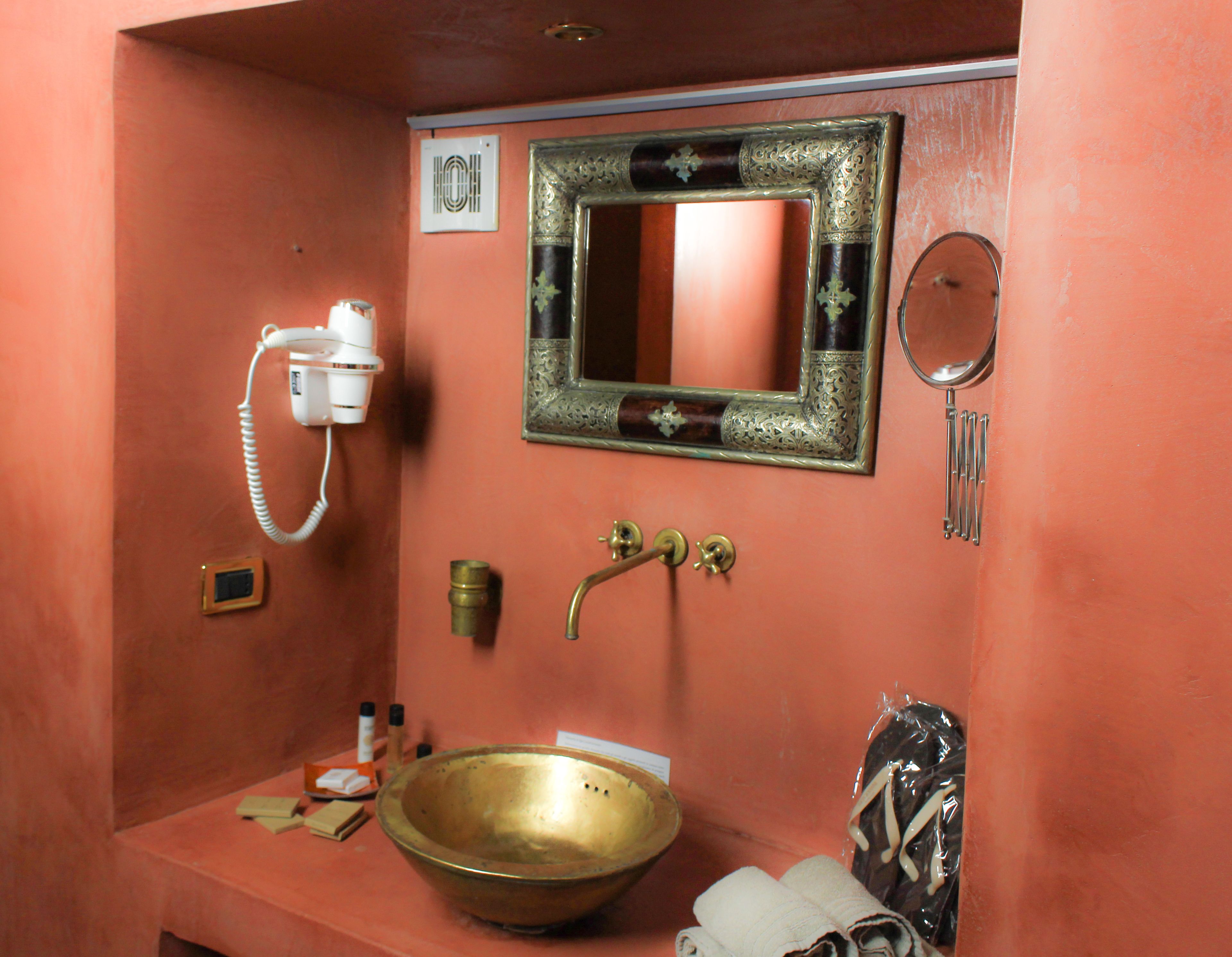 A bathroom with a brass sink, a mirror with an ornate frame, a hairdryer, and a small cabinet.