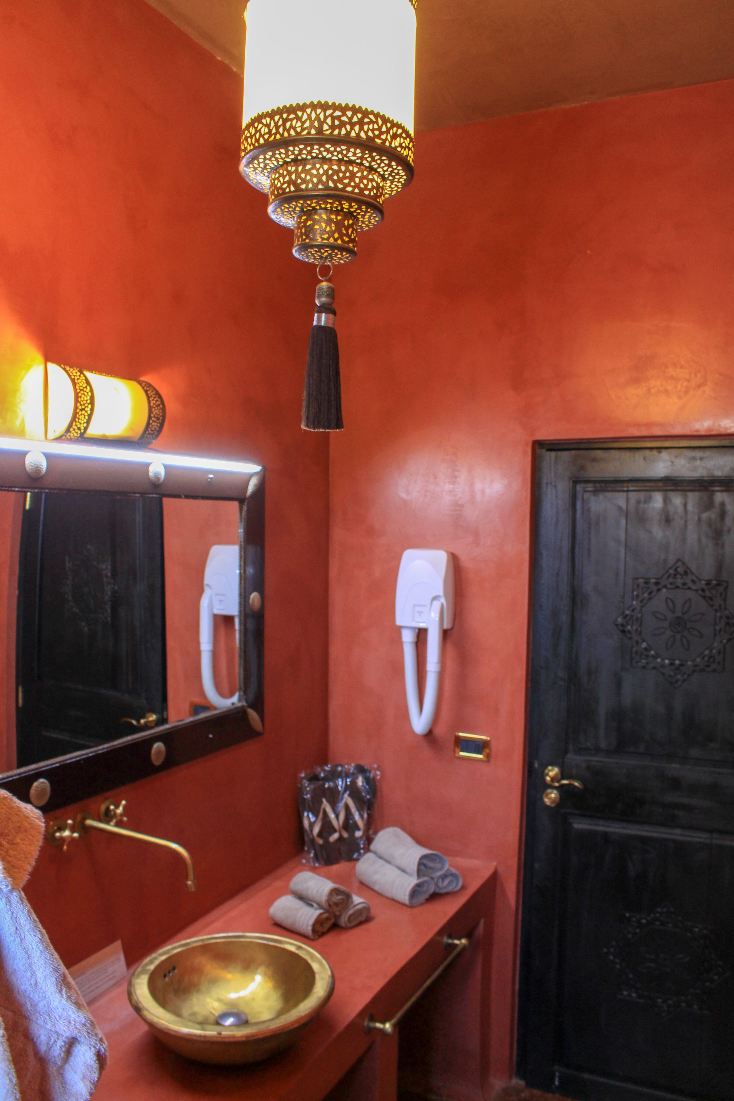 Un bagno con una parete rossa, un lavandino in ottone, uno specchio, un porta asciugamani, una porta con pannello decorativo e un lampadario.