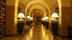 Un couloir aux plafonds voûtés et au sol orné de motifs, qui mène à une pièce où se trouvent un grand vase et une lampe.