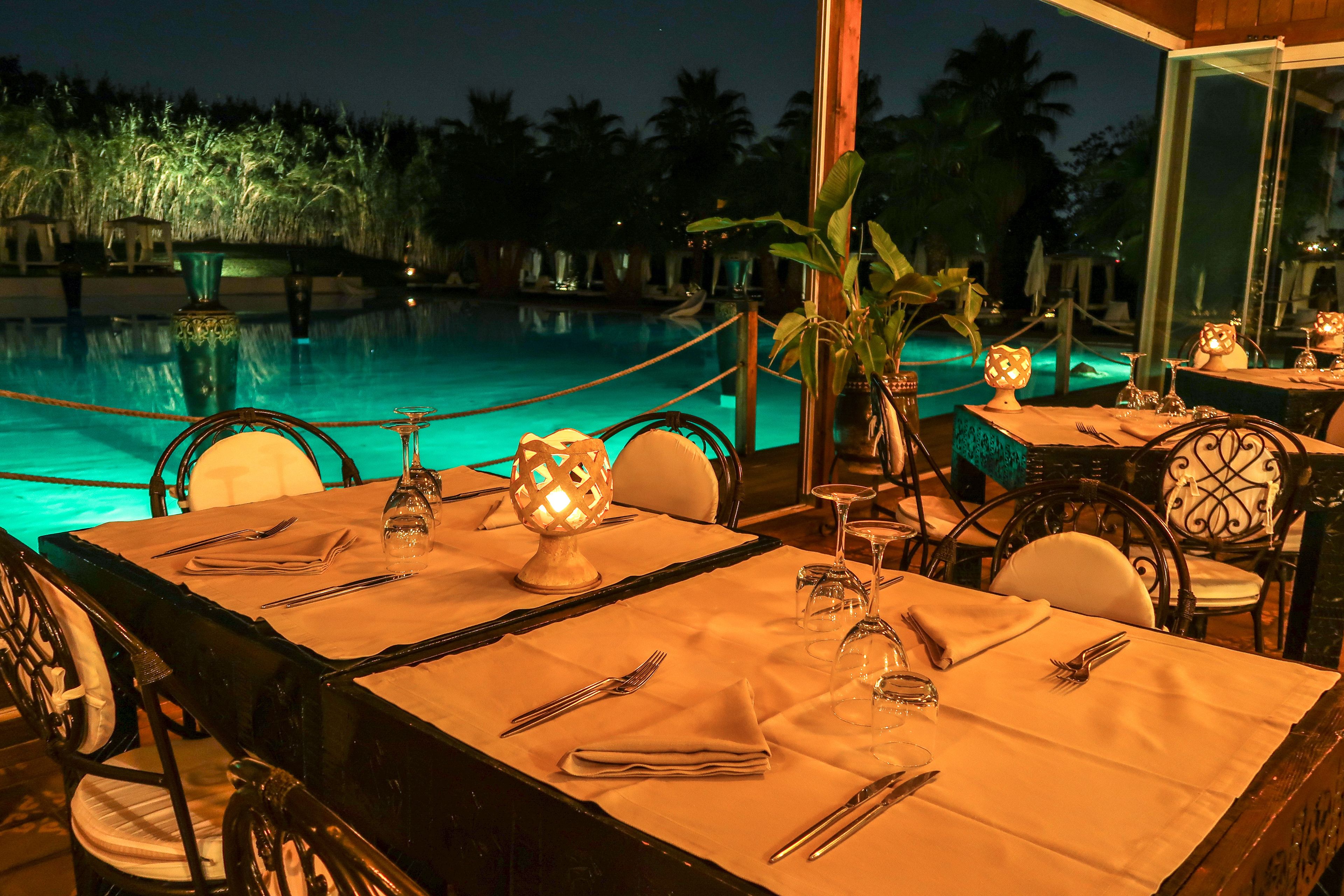 Ein Essbereich mit gedeckten Tischen, mit Blick auf einen Pool. Bei Nacht sind Palmen und eine beleuchtete Fontäne zu sehen.